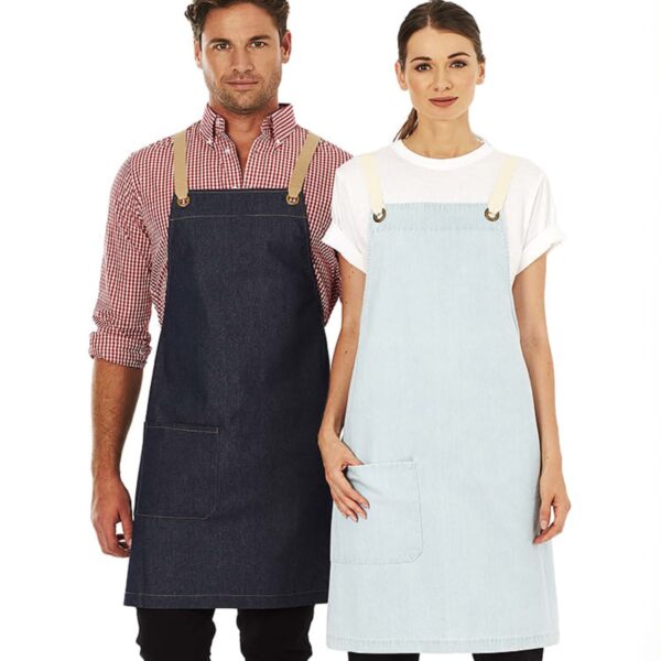 A man and a woman standing side by side, both wearing aprons. The man is in a dark denim apron over a red checkered shirt, and the woman is in a light denim apron over a white t-shirt. Both have hands resting at their sides.