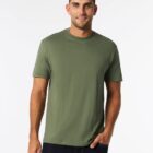 A person wearing a green t-shirt and dark jeans is standing against a plain white background. They have short dark hair and a slight smile, with one hand in their pocket.