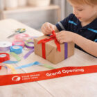 A child ties a red ribbon near Grand Opening Ribbons displayed on the table.