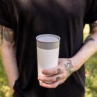 A person wearing a black T-shirt holds a white travel mug with a gray lid. Their arms have several tattoos. The background shows green grass, indicating an outdoor setting.