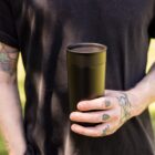 A person wearing a black T-shirt holds a black tumbler in their right hand. Their arm and hand have various colorful tattoos. The background is blurred with a green hue, suggesting an outdoor setting.