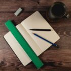 Open dotted notebook on a wooden table with a green ruler, black pen, blue pencil, eraser, and a mug of coffee.