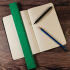 An open notebook with dotted pages lies on a wooden surface. A green ruler, a black pen, and a blue pencil are placed on the notebook.