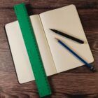 A dotted notebook lays open on a wooden surface with a green ruler, black marker, and blue pencil on top.