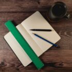 A dotted notebook is open on a wooden surface, with a green ruler, a black pen, and a blue pencil lying across it. A mug filled with a dark liquid is placed beside the notebook.
