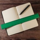 An open notebook with dotted pages lies on a wooden surface. A green ruler is placed diagonally across the pages, and a black pen rests on the notebook.