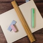 An open notebook with lined pages lies flat on a wooden surface. A wooden ruler is placed diagonally across it. To the left are two erasers, one pink and one blue, and to the right is a green pen.
