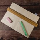 An open notebook with lined pages lies on a wooden surface. A wooden ruler is placed diagonally across the notebook. A pink eraser and a green pen are positioned beside the notebook.