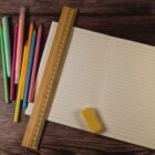 An open lined notebook lies on a wooden surface. A wooden ruler and a yellow eraser rest on the notebook. To the left, there are colorful pencils and pens scattered on the table.