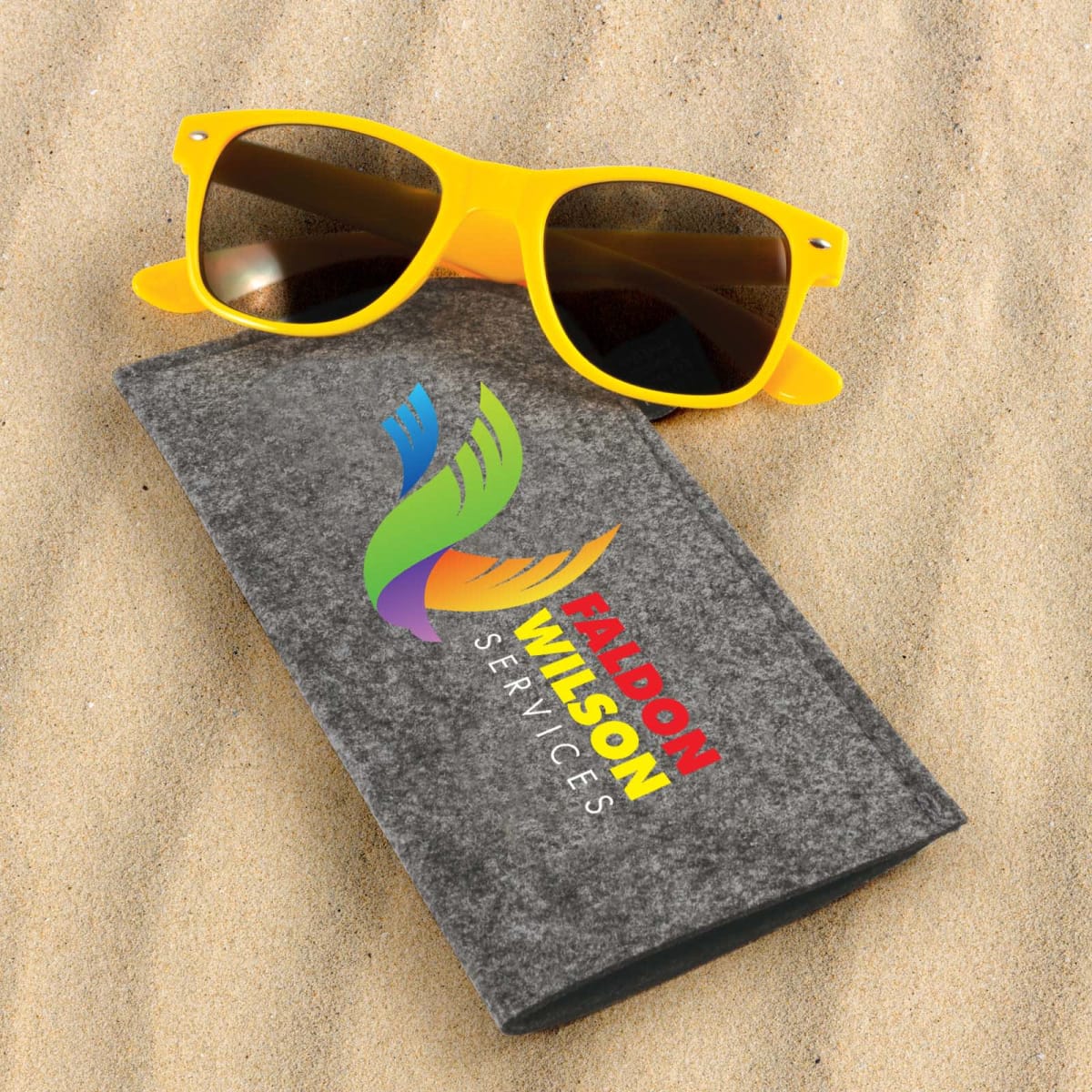 Yellow sunglasses and a gray box with a colorful logo and FALCON WILSON SERVICES text, resting on sand.