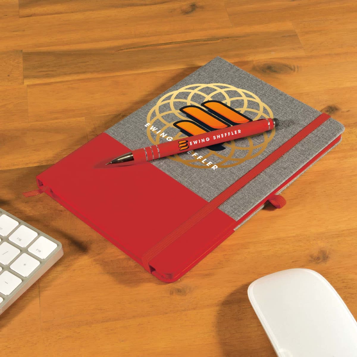 A red and gray notebook with a logo and the text Ewing Sheffler lies on a wooden table. A matching red pen is placed diagonally on top. A white keyboard and mouse are nearby.
