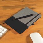 A closed gray and black notebook with an elastic strap, placed on a wooden desk. A black pen is resting on it. A section of a white computer keyboard and a wireless mouse are visible in the foreground.