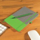 A closed notebook with a green and gray cover lies on a wooden desk. A green pen is placed on top of the notebook. Part of a white keyboard and a white mouse are visible at the edges of the image.
