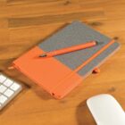 A notebook with an orange and gray cover lies on a wooden desk. An orange pen rests on top of it. Nearby are the corner of a white keyboard and a white computer mouse.