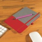 A notebook with a gray and red cover lies closed on a wooden desk. A pink pen is placed diagonally across it. Part of a keyboard and a white mouse are visible on the desk.