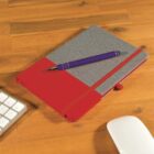 A closed notebook with a red and gray cover lies on a wooden desk. A purple pen is placed diagonally on top of it. Nearby, a section of a white keyboard and a white computer mouse are partially visible.