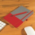 A red and gray notebook with a red pen is placed on a wooden desk. A white keyboard and a white computer mouse are partially visible in the background. The notebook has a red elastic band and matching ribbon bookmark.