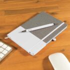 A closed, gray and white notebook with a white pen rests on a wooden desk. Beside it, part of a white computer keyboard and a white computer mouse are visible. The notebook has an elastic band and a pen loop.