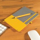 A notebook with a gray and yellow cover lies on a wooden desk. A yellow pen is placed diagonally on top of the notebook. Part of a white computer keyboard and a white wireless mouse are visible nearby.