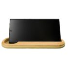 A smartphone placed horizontally on a wooden charging stand. The phones screen is off, and the stand has a light brown finish with a groove to hold the device.