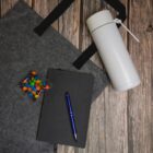 Flat lay of a notebook, pen, candies, tote bag, and a white flask on a wooden surface.