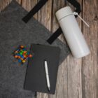 Tote bag, thermos flask, notebook, pen, and candy on a wooden surface.