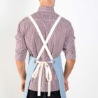 Man in a checkered shirt wearing a blue apron with crossed white straps on the back and tied at the waist, standing against a plain white background.