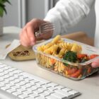 A person eating pasta with vegetables from a plastic container near a computer keyboard. The container has a partially removed lid labeled Roll Done. A hand holds a fork above the meal. A small plant is visible in the background.
