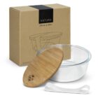 A clear glass food container with a bamboo lid is displayed in front of a brown cardboard box labeled Natura. A small white strap is beside the container. The box features an illustration of the container and lid.
