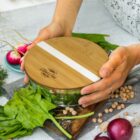 A person places a bamboo lid on a glass salad bowl filled with greens. The lid has a logo and white stripe. Surrounding are radishes, spinach leaves, chickpeas, and a red onion on a textured surface.
