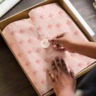 A person is sealing a box with branded pink tissue paper using a round sticker. The paper and sticker both feature a logo with the letters Be in a cursive font. The box is placed on a dark wooden surface.
