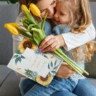 A woman and child share a warm embrace on a couch. The woman holds a bouquet of yellow tulips and a card decorated with sunflowers and leaves. The card has an affectionate message on the front.