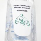A clear hand sanitizer bottle with a carabiner clip attached. The label reads Logan Community Visitors Scheme with the phone number 3290 0088. An abstract design resembling a stylized heart or lungs is also on the label.