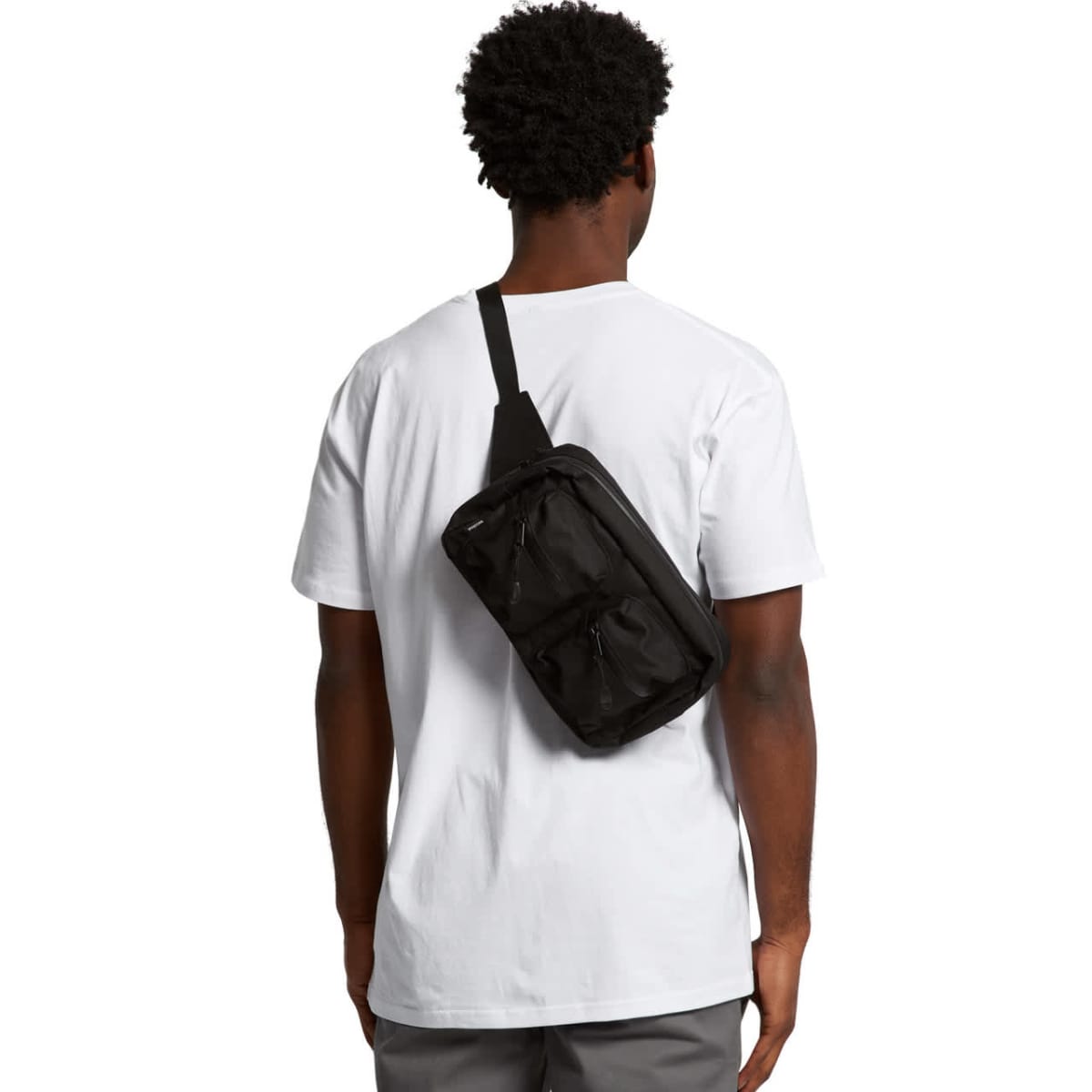 A person with curly hair stands facing away, wearing a plain white T-shirt and gray pants. They have a black crossbody bag slung over their shoulder. The background is plain white.