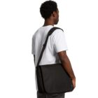 A person wearing a white t-shirt is seen from behind carrying a large black messenger bag over their shoulder. The person has short curly hair and is standing against a plain white background.
