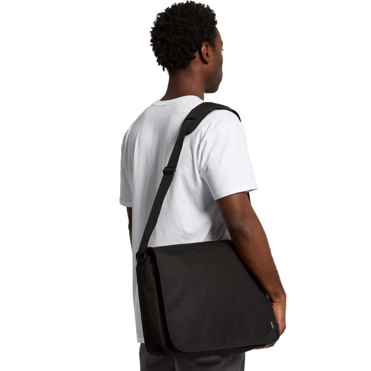 A person wearing a white t-shirt and gray pants stands with their back to the camera, holding a large black messenger bag over their shoulder. Their head is slightly turned to the side.