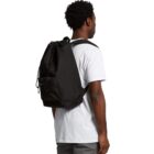 A person with short curly hair is wearing a white T-shirt and gray pants, carrying a large black backpack. The person is facing away from the camera.
