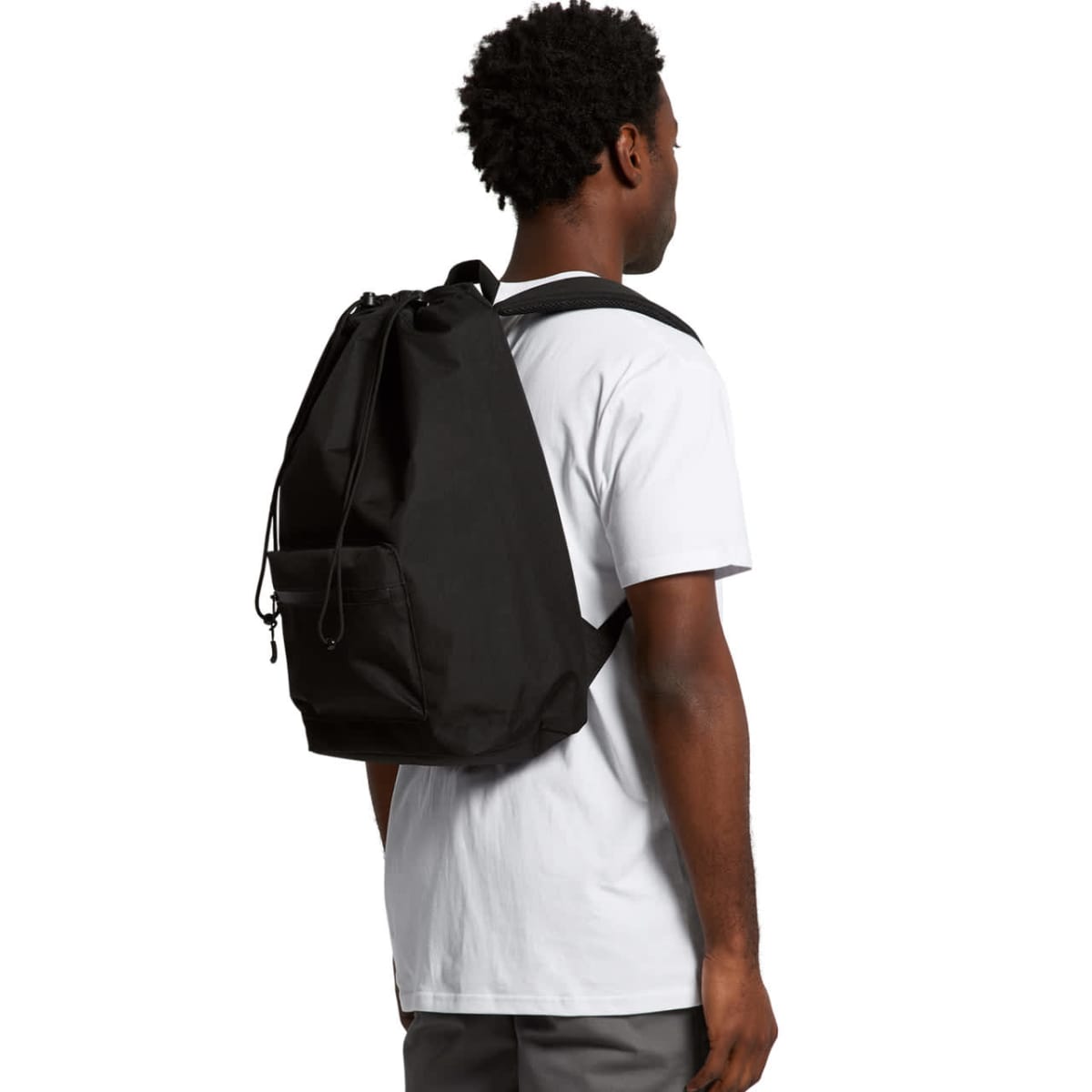 A person wearing a white T-shirt is standing with their back to the camera, carrying a large black backpack. The person has short curly hair and is wearing gray pants.