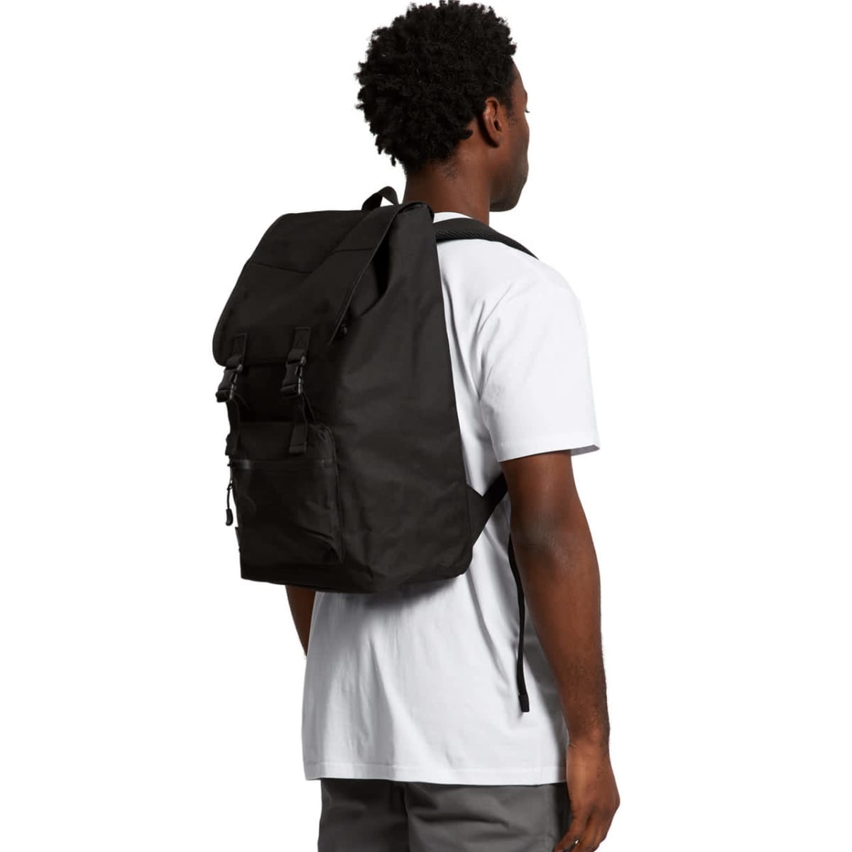 A person with short curly hair, wearing a white T-shirt and gray pants, is standing and facing away from the camera. They are carrying a large black backpack.