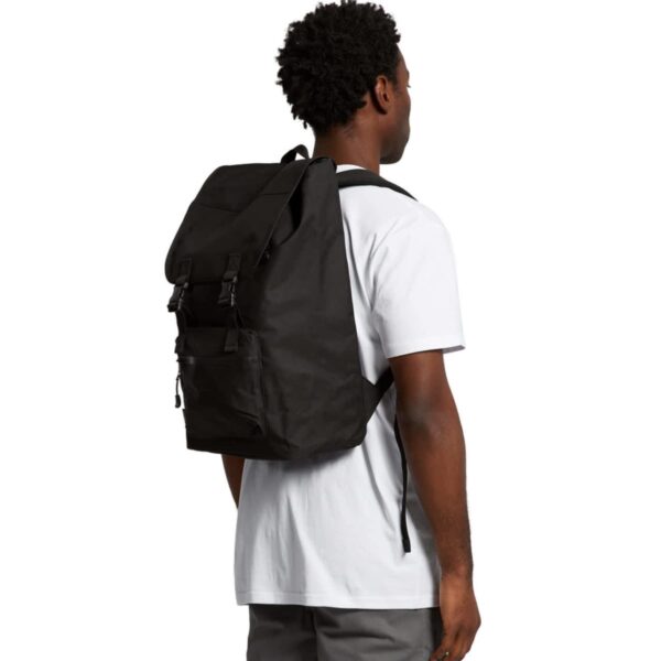 A person wearing a plain white t-shirt and gray pants stands with their back to the camera, carrying a large black backpack. The backpack has a fold-over top secured with straps and buckles. The person has short, curly hair.