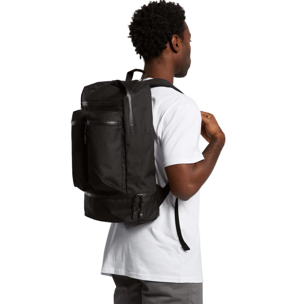 A man wearing a white t-shirt and gray pants, carrying a large black backpack. He is facing slightly away, looking to his left.
