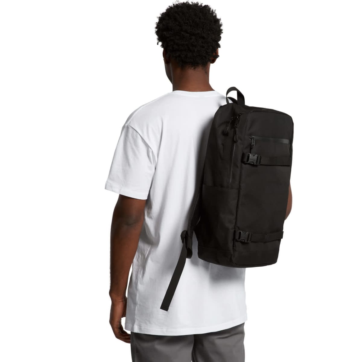A person with short curly hair is wearing a white T-shirt and gray pants, carrying a large black backpack over one shoulder. The individual is facing away from the camera.