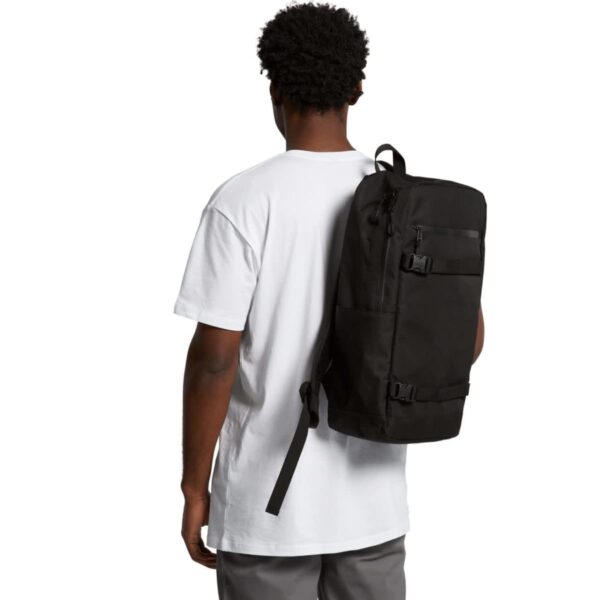 A person wearing a white T-shirt and gray pants is carrying a large black backpack over one shoulder. They are facing away from the camera.