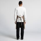 Rear view of a person standing with their arms at their sides, wearing a white long-sleeve shirt, black pants, and a brown apron tied at the back. The background is plain and light-colored.