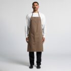 A person wearing a long brown apron stands facing forward. They have a white long-sleeve shirt and black pants with black shoes. The background is plain gray.