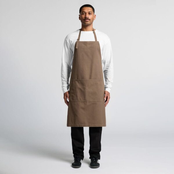 A person stands against a plain background wearing a brown apron over a white long-sleeve shirt and black pants. They have short hair and a mustache, looking directly at the camera with a neutral expression.