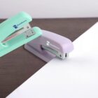 Two staplers, one mint green and the other lavender, are positioned on a wooden surface. They face a blank sheet of white paper, ready to be used. The staplers have a small rectangular logo with unreadable text.