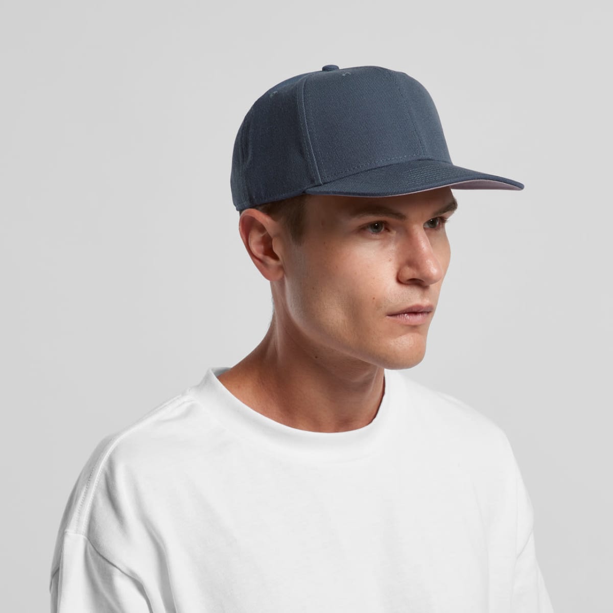 A person wearing a dark blue baseball cap and a white T-shirt is looking to the side against a plain gray background.