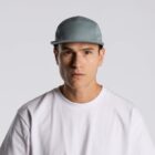 A person wearing a light grey cap and a plain white T-shirt against a grey background. Their expression is neutral.