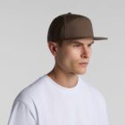 A person wearing a brown flat-brimmed cap and a white t-shirt stands against a plain gray background, looking to the side.